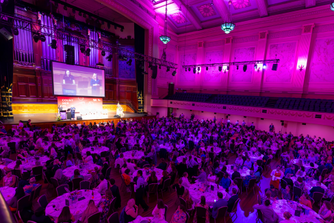 A birds eye view of a room of 700 people sitting at round tables of 10. There's purple lighting. 