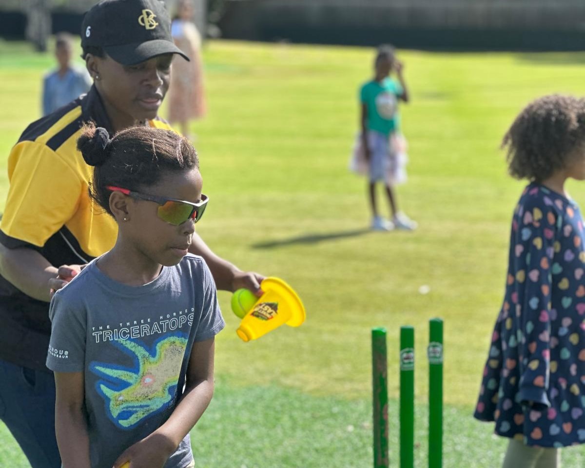 A Zimbabwean cricketer is coaching a young girl next to a cricket wicket on an oval. There are 4 other kids in the background of the image, who are out of focus. 