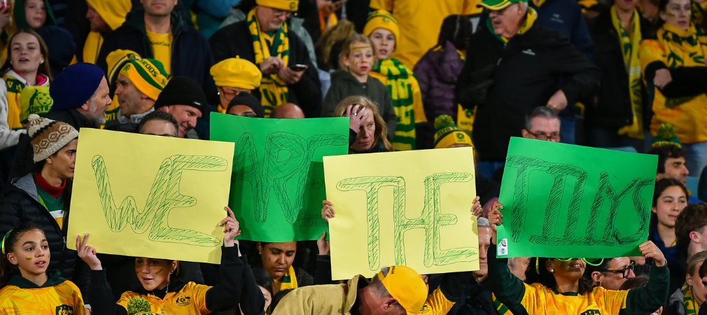 Fans in the stadium holding up bright yellow and green signs that spell out 'WE ARE THE TILLYS,' surrounded by a crowd dressed in Australian colours, showing their support for the team. 