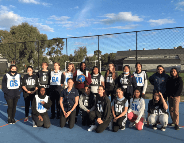 a group photo of a female netball team standing on an outdoor netball court. they are wearing their netball bibs showing their positions, smiling at the camera. 