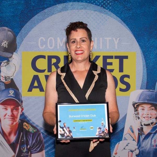 A woman stands proudly with a certificate saying Congraulations Burwood Cricket Club, Community Parnership. She stands in front of a colourful banner with lifesize images of young female cricketers on it. 