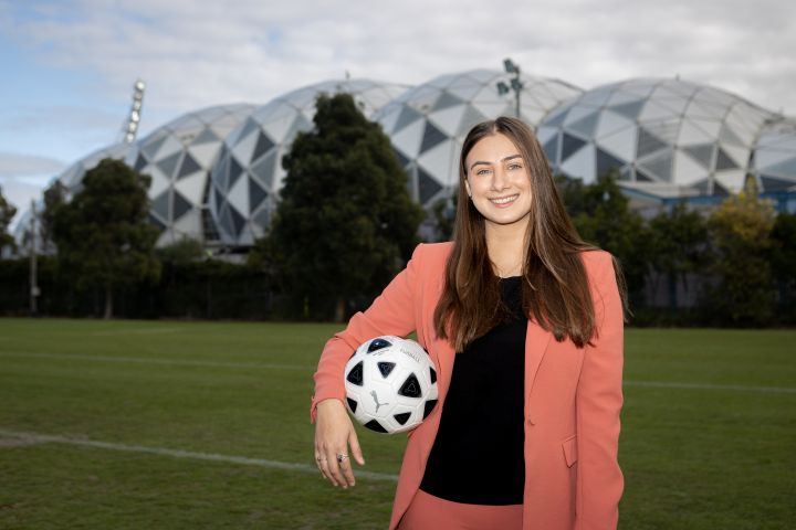 Successful Women in Sports Broadcasting applicant, Georgia Rajic, standing with a soccer ball under her arm smiling at the camera